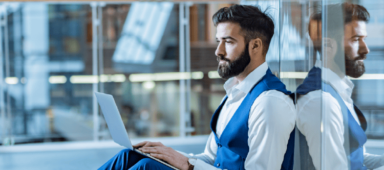 Homem com barba usando terno azul, concentrado em seu laptop em ambiente moderno. Ideal para ilustrar produtividade e trabalho.
