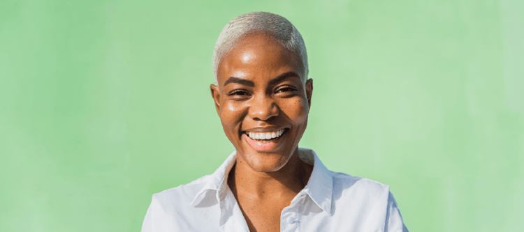 Mulher sorridente com cabelo curto e loiro, vestindo uma camisa branca, em frente a um fundo verde vibrante. Representa confiança e alegria.