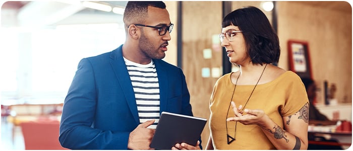 Duas pessoas em um escritório conversam, uma mulher gesticula enquanto um homem segura um tablet, ambos focados na interação.