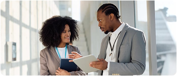 Duas pessoas, conversam em um ambiente iluminado, com um homem segurando um tablet e uma mulher sorrindo para ele.