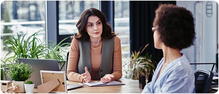 Duas mulheres em um escritório conversam em uma mesa, com uma gesticulando enquanto a outra a ouve atentamente.